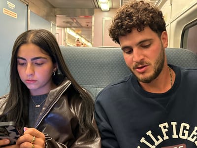 The scene depicts two young adult individuals seated inside a commuter train car. The location is a train operating within the city of East Rutherford, United States.

On the left, a female subject with long, dark hair parted in the middle, and dark eyebrows, is shown. She is wearing a dark brown faux leather jacket over a grey crew-neck sweater. A light-colored pendant necklace is visible around her neck, along with silver hoop earrings. Gold-toned rings are present on her right hand. She is holding a smartphone with a dark patterned case in both hands, looking down at its screen.

On the right, a male subject with curly brown hair and a short beard is seated. He is wearing a navy blue crew-neck sweatshirt featuring a white "HILFIGER" logo across the chest. A thin, dark beaded necklace is visible around his neck. His gaze is directed downwards and slightly to his left, and his mouth is slightly ajar.

The interior of the train car features blue and grey upholstered seats, light blue wall panels, and overhead fluorescent lighting fixtures. An "EMERGENCY RELEASE" sticker with red text is visible on a wall panel in the upper left background. In the reflection of a doorway or window at the far end of the car, two blurry figures are discernible, one wearing a light-colored baseball cap backward. The subjects are seated next to each other; the female is actively using her phone, while the male appears to be observing something or listening.