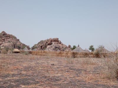 The image depicts a rural scene in Shahid Afandi, Sudan. The foreground consists of dry, barren land covered with straw and scorched patches. In the midground, a low, woven fence encloses a village of huts with thatched roofs. Rocky hills and sparse vegetation define the background. There are a few human figures in the image, possibly inhabitants of the village. The overall scene suggests an arid environment and a traditional way of life.