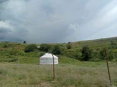 A wide shot captures a yurt, a traditional circular dwelling, situated in a grassy, rolling landscape under a heavily overcast sky. The yurt is predominantly white with a decorative red and white patterned band around its lower section. It is positioned in the middle ground, behind a wire fence that runs horizontally across the foreground. The fence is supported by metal posts. The terrain surrounding the yurt is uneven and covered in green grass and scattered bushes. In the distance, on the far right, a small, dark-colored structure is visible atop a hill. The sky is filled with dark, grey clouds, suggesting an impending storm or recent rain. A single, bare-branched tree stands on a distant hill to the left. The scene appears to be in a rural or semi-rural area, likely the region of Көксаяқ ауылдық округі, Kazakhstan, characterized by its open terrain and traditional dwellings. There are no people visible in the image.
