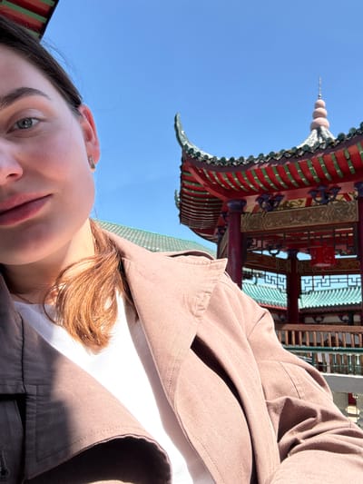 A person is captured in a low-angle selfie, with part of her face, brown hair, and shoulder visible on the left and lower right of the frame. She has fair skin, wears a small ear piercing, and is dressed in a light brown or taupe jacket over a white top. The image is taken outdoors on a bright, sunny day, indicated by the clear blue sky and strong daylight.
In the background, dominating the upper and right portions of the frame, is a vibrant building with distinct East Asian architectural features. The structure showcases a multi-tiered roof adorned with red and green tiles or decorative elements, intricate wooden beams, and latticework. A slender finial or spire rises from the top of the roof against the sky.
The scene suggests a moment of leisure, possibly a tourist or visitor taking a picture with the decorative architecture. Given the provided location context of Hainburg an der Donau, Austria, this building is likely an East Asian-style temple or pagoda situated within that area, such as the Hainburg Pagoda or Buddhist Center. No visible text is present in the image.