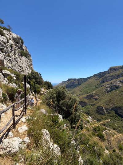 The image depicts a scenic hiking trail with two individuals. The trail is carved into a rocky, mountainous landscape with sparse vegetation. A wooden railing lines the left side of the path. A woman in a hat and light-colored clothing is walking on the trail, looking towards the camera and gesturing with her hand. Behind her, a man in a blue shirt and hat is also walking on the trail. The background shows a vast valley with steep, green-covered slopes and rocky outcrops, leading to a distant view of the sea under a clear blue sky. This location is likely in the Vendicari Nature Reserve near Noto, Italy, known for its rugged coastal trails and natural beauty.