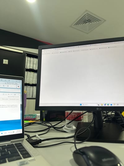 The image captures an office workspace, seen from a high angle looking down and slightly to the right, in Sanctorum, Mexico. The primary subjects are two computer screens: a large LG monitor dominating the left side, and a laptop screen visible on the right.

The LG monitor displays a mostly white web browser interface with numerous open tabs. Visible tab titles include "reforma de pension" (pension reform), "End of Trials (13).xlsx," "NetScaler Gateway," "EDAG Mexico - Car...", "Feed | Linkedin," "Vacantes_PD_IE_2026.xlsx" (Job vacancies_PD_IE_2026.xlsx), "Contraseñas.xlsx" (Passwords.xlsx), and "Rastreo-DHL-México" (Tracking-DHL-Mexico). A search bar with a magnifying glass icon is at the bottom center of the screen, and application icons like Teams and Outlook are arranged vertically on the right edge, next to the "LG" logo.

To the right, a silver laptop with a dark keyboard is open, displaying an email. The email subject reads "Seg. Reclu EoD II - Rocio," with a note about "Tomorrow 11:30 AM Micros.....". The email's timestamp is "Wed 4/29/2026 10:22 AM," and a snippet of the body reads, "AG Mexico. If your qualifications are about your fit in this position." The laptop's system time shows "3:09 PM" and the date "4/29/2026," with "ENG" indicating the English language setting.

Various desk items are scattered between and around the monitors. A black computer mouse is partially visible on the far right. A red cylindrical container labeled "Clips" with Spanish text "orrados de vinil" (likely "vinyl clips") sits near the LG monitor stand. Multiple black and white cables are tangled behind the monitors, connecting the devices. A white charging cable, a purple pen, and a small green/blue object (possibly a stapler) are also visible.

In the background, behind the screens, several shelving units filled with what appear to be binders or files are neatly arranged. A white square air conditioning vent is prominent on the ceiling in the upper left corner of the image. The time displayed on the laptop is 3:09 PM, on what appears to be an ordinary workday, given the office setting and content on the screens.