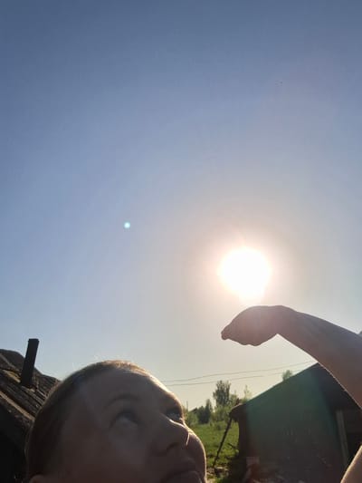 A selfie oriented in a way that suggests the photograph was taken looking upward towards the sky. A person's face is visible in the top left corner of the image. An arm is extended toward the sun, which appears as a bright white circle in the lower center of the image. The sky is a clear gradient of blue, becoming lighter towards the horizon. The lower left corner also contains a part of what appears to be a green-colored roof. The location is Шешурга, Russia.