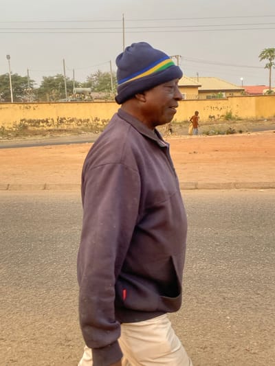 An outdoor, eye-level shot captures a man standing on what appears to be a street or roadside in Basawa, Nigeria. He is positioned in the foreground, facing right with his head tilted slightly upwards, looking towards the hazy sky. He is wearing a dark blue long-sleeved collared shirt, light-colored trousers (possibly white or off-white), and a dark blue beanie adorned with yellow and light green horizontal stripes. A small, indistinct red tag is visible on the lower left side of his shirt.

The background reveals a simple street environment. To the right of the asphalt road where the man stands, there's an unpaved, earthy shoulder bordered by a light brown or tan wall. Another individual is visible in the mid-ground, walking or standing on this unpaved strip further down the road, facing away from the viewer. The sky is bright but noticeably hazy or dusty. Utility poles with power lines run across the upper part of the frame, and sparse green trees are scattered throughout the background. Buildings with light-colored walls and simple designs can be seen in the distance. The lighting indicates it is daytime, likely late morning or afternoon. No discernible text is visible in the image.