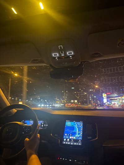 The image depicts the interior of a car driving at night through what appears to be a busy urban environment. Based on the visible skyline and building styles, it is possible that the location is Zeytinburnu, Turkey. Traffic is visible through the front windshield, and various dashboard elements are illuminated, including the navigation screen and steering wheel controls. A hand is seen interacting with the steering wheel. The interior of the car appears relatively clean.