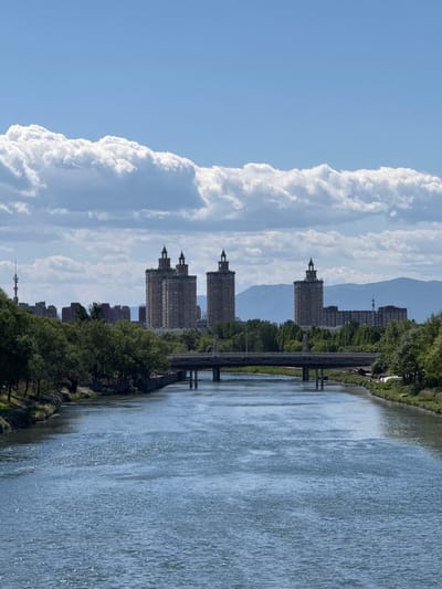 The image shows a river scene in Changping District, China. The river reflects the buildings and sky. Buildings line the riverbank. Trees and other vegetation are present. The sky is blue with large, billowing white clouds. A bridge spans the river. Mountains are visible in the distance. The photo is taken from a high angle.