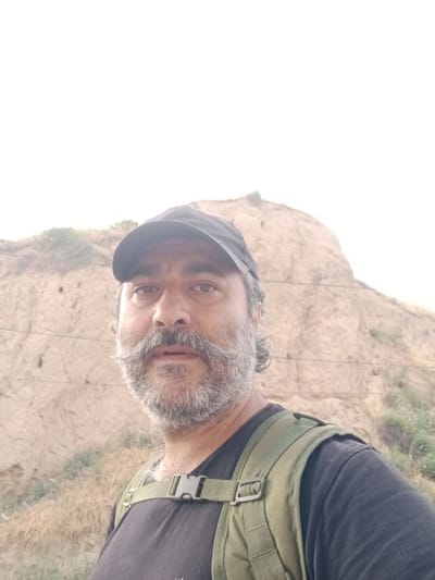 A close-up, waist-up, medium shot shows a middle-aged man with a grey beard and mustache, wearing a black baseball cap and a black t-shirt with a green backpack. He is looking directly at the camera. The background is a blurred, tan-colored hillside under a bright sky. The scene appears to be outdoors. The location is identified as Parkent, Uzbekistan.