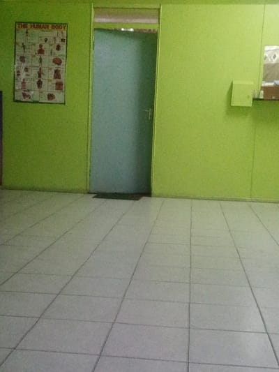 The image depicts the interior of a room, likely a medical facility or office waiting area, located in Majengo ward, Kenya. The walls are uniformly painted a bright lime green. The floor is covered with light grey square tiles laid in a consistent grid pattern.

Centrally positioned on the rear wall is a closed, light blue-green door, which appears to be made of frosted glass or a similar opaque material. The door has a silver-colored horizontal handle. A dark, rectangular doormat is situated directly in front of the door at floor level. Above the door, a narrow transom window provides a glimpse of an adjacent, slightly lighter area.

On the left green wall, a white poster titled "THE HUMAN BODY" is affixed. This poster displays various anatomical diagrams of human organs and systems. On the right green wall, a small, square, light green wall-mounted cabinet or box is visible. To the right of this cabinet, a reflective surface, possibly a mirror or tiled section with a diamond pattern, is situated above what appears to be a counter or shelf.

No individuals are present within the scene, and no actions or interactions are depicted. The environment appears clean and well-lit.