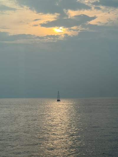 A single sailboat with a tall mast is centered on a calm ocean. The sun is setting behind a bank of clouds, casting a warm, golden light on the water. The sky is overcast, with patches of blue sky visible through breaks in the clouds. The scene is serene and peaceful. The location is Unknown, Malaysia.