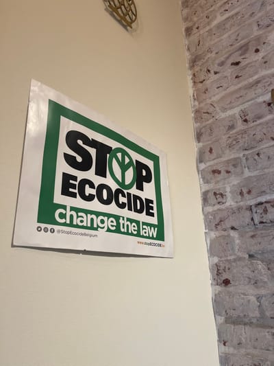 An indoor scene in Saint-Gilles - Sint-Gillis, Belgium, features a white poster with a green border affixed to a light-colored painted wall. The poster prominently displays "STOP ECOCIDE" in large black capital letters, with the 'O' in 'STOP' replaced by a green stylized peace symbol resembling a plant or tree. Below this, "change the law" is printed in smaller black text. The bottom of the poster includes social media icons (Instagram, Facebook) with the handle "@StopEcocideBelgium" and the website address "www.stopECOCIDE.be". The poster shows a slight curvature along its bottom edge. To the right of the painted wall, an exposed brick wall with light grey and reddish-brown bricks and white mortar forms an interior corner. A golden-colored, ornate, circular metal decoration is partially visible on the upper section of the painted wall. No individuals are depicted.