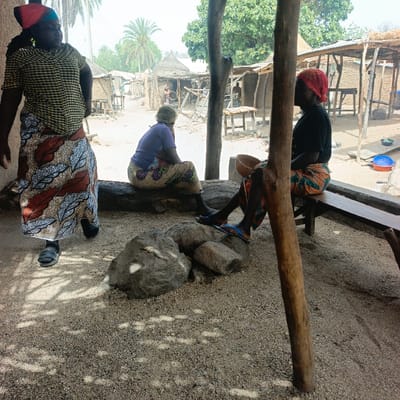 The image displays an outdoor scene in Boi B, Nigeria, featuring three adult females and traditional village infrastructure. The foreground is composed of light brown, sandy-gravelly soil, partially shaded by an unseen structure, creating dappled sunlight patterns. Two prominent, thick, rustic wooden posts are visible; one on the far left, another vertically in the mid-right.
On the left, a female subject stands in motion, angled slightly away from the camera. She wears a yellow and black checkered short-sleeve top, a long skirt patterned with white, brown, black, and orange leaf motifs, and a red headscarf. Dark sandals are on her feet.
In the center, a second female subject is seated on a large, horizontal wooden log, facing away from the camera and looking towards the background. She wears a light purple short-sleeve top, a multi-patterned wrapper or skirt, and a green head covering.
To the right, a third female subject is seated on a simple wooden bench, facing left. She wears a dark-colored short-sleeve top, an orange, brown, and green patterned wrapper or skirt, and a red headscarf. She wears blue flip-flops with lighter blue straps and holds a light brown, wooden bowl in her lap.
A cluster of three to four grey rocks is positioned on the ground in the foreground, between the standing and seated subjects.
The background reveals a village pathway lined with multiple rustic structures constructed from mud, wood, and thatch roofs, consistent with huts or open market stalls. Palm trees and other green foliage are visible beyond these structures. In the far right background, a blue plastic bowl and a red plastic bowl are present on the ground. The background is brightly lit by natural sunlight, indicating an outdoor environment.
