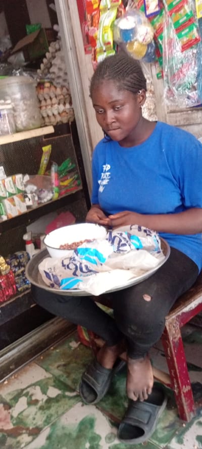 In Kwachiri, Nigeria, a young Black female sits on a red stool with a metal tray on her lap. She is wearing a blue t-shirt with white lettering and black leggings. On the tray, there is a white bowl containing brown beans and rice, and a plastic bag with blue and white patterns. Her feet are bare, and she is wearing dark-colored sandals. The background shows shelves stocked with various food items, including eggs and packaged snacks, suggesting a market or store setting. The floor is tiled with a green and white pattern.