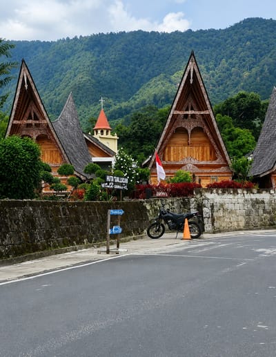 The image captures a scenic view in Ambarita, Indonesia, featuring traditional Batak architecture set against a lush, forested mountain backdrop. In the foreground, a paved road runs alongside a low stone wall. A black motorcycle is parked on the sidewalk next to the wall, with an orange traffic cone beside it. Two blue directional signs are mounted on wooden posts near the motorcycle, indicating "PARKING" to the left and an arrow pointing to the right. Behind the wall, several wooden houses with distinctive triangular roofs, adorned with intricate carvings, are visible. The Indonesian flag is displayed on one of the houses. A church steeple with a red roof and a white cross rises above the houses. The surrounding area is filled with greenery, including trees and red foliage. The sky is partly cloudy, and the mountains in the distance are covered in dense forest.