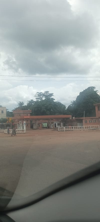 The image depicts a Total gas station in Ozalla, Nigeria. The station is located on a dirt road, and there are a few people standing near the pumps. The sky is overcast, and there are trees in the background. A motorcycle is present on the left side of the image, near the gas station. White metal barriers are positioned in front of the station. A vehicle interior is visible in the foreground, suggesting the image was taken from inside a car.