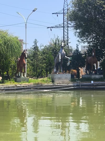 The image depicts a serene outdoor scene, likely in the city of Vahdat, Tajikistan, dominated by a calm, green body of water in the foreground. The water reflects the surrounding environment, including trees and structures.
In the background, three prominent statues of horses are displayed. The leftmost statue is a brown horse standing with all four legs on a pedestal. To its right, a light gray horse is depicted in a rearing posture, with its front legs raised. The rightmost statue is another brown horse, also standing on a pedestal.
Behind the statues, lush green trees and vegetation create a natural backdrop. A large, intricate electrical pylon stands tall, suggesting the presence of power infrastructure in the vicinity. A street lamp with two lights curves gracefully on its pole. In the distance, buildings are visible, hinting at an urban or semi-urban setting. The overall atmosphere is tranquil and picturesque, bathed in bright daylight under a clear blue sky.