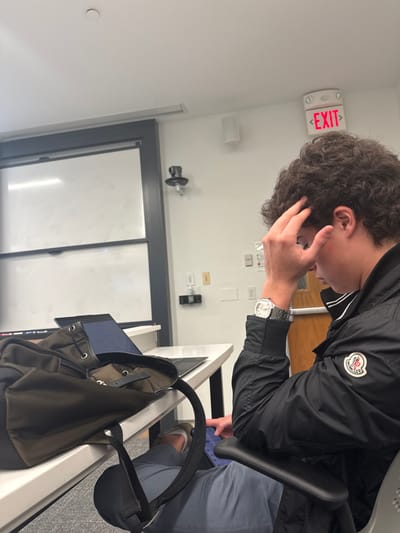 An indoor scene in a modern classroom or study environment in Wellesley, United States, captures a young adult male seated at a white desk. The male, positioned on the right side of the frame, has dark, curly hair and wears a black zippered jacket with a white and red circular Moncler logo on the left bicep, grey/blue trousers, and brown shoes. A silver-toned wristwatch is visible on his left wrist. His right hand is raised, fingers touching his forehead/temple area, as he looks downwards, possibly at a tablet or small device on his lap. On the white desk in front of him, a black laptop is open with a lit screen displaying text. A dark green/brown backpack rests to the left of the laptop. The background features a large white projection screen or whiteboard with a dark grey frame on the left. A black security camera or sensor is mounted on the white wall above waist height. An illuminated red "EXIT" sign is affixed to the ceiling in the upper right. White wall-mounted speakers and various electrical outlets are also present on the white wall. A portion of a wooden door frame is visible on the far right.