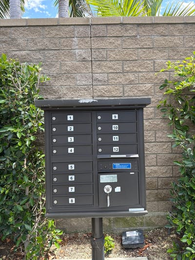 A dark gray, multi-unit mailbox stand is affixed to a textured tan brick wall. The stand is oriented vertically and features two rows of mailboxes. The top row has mailboxes numbered 9 through 12, while the bottom row displays mailboxes numbered 1 through 8. Each mailbox has a small, circular knob or latch at the bottom. Mailbox 12 has a blue and white sticker indicating "USPS OUTGOING MAIL DEPOSIT MAIL FOR COLLECTION". There's also a silver lock and handle on one of the mailboxes, suggesting it's a common mail delivery point.
To the left of the mailbox stand, a large palm tree with green fronds is visible against a bright blue sky with some white clouds. To the right and below the mailboxes, lush green bushes and foliage surround the base. The ground is covered with dirt, small rocks, and fallen leaves. There's also a black plastic receptacle with some writing on it located to the right of the mailboxes. The lighting suggests it is daytime, likely sunny, given the shadows and bright foliage. The setting appears to be an outdoor area, possibly a residential complex or public space in Irvine, USA, given the context. No people are visible in the image.