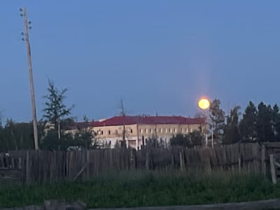 The image depicts a rural scene in Khorobut, Russia, at dusk. In the foreground, a weathered wooden fence runs horizontally across the frame, partially obscured by tall green grass. Beyond the fence, a long, light-colored building with a red roof is visible, featuring numerous windows. Trees surround the building, with some appearing denser and darker on the right side of the frame. A tall wooden utility pole stands to the left of the building, supporting wires that stretch across the clear blue sky.
The most striking element in the sky is a large, luminous full moon, casting a warm, yellowish glow. The moon is positioned above and slightly to the right of the building, appearing prominent against the twilight sky. The overall atmosphere suggests a quiet, serene evening in a rural setting. There is no visible text in the image.