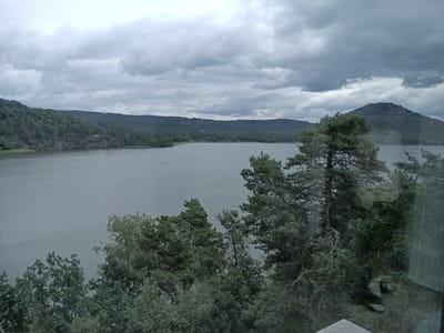 The image shows a scenic view of a lake surrounded by lush green trees and hills, under a cloudy sky. The water is calm, reflecting the overcast sky. In the foreground, dense foliage of trees partially obscures the view, with a glimpse of a wooden structure at the bottom right corner. The background features a long stretch of tree-covered hills, with a prominent rounded hill on the right side, topped with a structure that appears to be a tower or antenna. A small boat is visible on the water in the distance. This vista is characteristic of the landscape around Lake Mácha (Máchovo jezero) in Doksy, Czechia, a popular tourist destination known for its natural beauty. The overall impression is one of serene, natural beauty on a subdued, cloudy day.