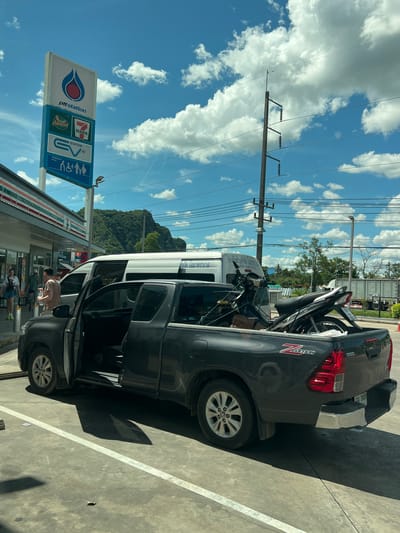 The image shows a dark gray Toyota Hilux pickup truck parked in what appears to be a gas station lot. The truck's driver-side door is open, and a motorcycle is loaded in the truck bed. Two other motorcycles are also visible in the truck bed, secured with straps. A white van is parked behind the pickup truck. To the left, a 7-Eleven convenience store is visible, with its distinctive green and red signage. A large sign for "ptt station" is also present, featuring the PTT logo and various other service indicators like "Amazon", "7-Eleven", and "EV STATION". Several people are walking near the convenience store. In the background, there are power lines and a lush green mountainside. The scene is illuminated by bright sunlight, casting shadows on the concrete ground, which is marked with white lines. The license plate of the pickup truck reads "UM 3409". The scene is located in Tambon Phraek Ha, Thailand, as indicated by the overall context of the brands and the likely geographical setting.