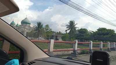 The image captures a view from a vehicle traveling through Cotpuuk, Indonesia, under a cloudy sky. In the distance, a mosque with distinctive teal-colored domes and minarets is visible. To the right of the mosque, palm trees and lush green vegetation surround a small, white building. A red-railed bridge or barrier runs horizontally across the foreground, with concrete posts supporting the structure. Overhead, several black electrical wires stretch across the frame. The interior of the vehicle is partially visible, with a tan-colored dashboard and part of a black phone mount in the lower right corner.
