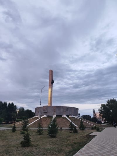 This image captures an outdoor scene featuring a large, concrete monument. The monument is characterized by a wide, circular base elevated on a grassy mound, with a tall, rectangular pillar rising from its center. The pillar is illuminated by a warm light, highlighting a dark object attached to its side. The monument is surrounded by a paved walkway and sparse evergreen trees. In the background, a line of trees and some buildings are visible. The sky is overcast with dramatic clouds, suggesting either early morning or late evening. The overall mood is somewhat somber due to the cloudy weather and muted colors, but the illuminated monument adds a focal point of interest. A single person is visible on the far right of the image, walking away from the monument. The setting appears to be a public space, possibly a park or memorial area. There is no visible text in the image.