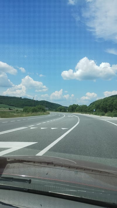 The image shows a paved road with white lane markings, curving through a landscape of green hills covered in trees and vegetation. The sky is blue with scattered white clouds. The perspective is from inside a vehicle, looking out through the windshield. The bottom of the image shows part of the vehicle's dashboard and windshield wipers. There is a subtle texture overlaying the image, giving it a vintage or artistic appearance. The scene is a natural, outdoor environment with no visible people or buildings.