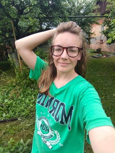 A young woman with blonde hair and glasses is taking a selfie in a grassy, overgrown backyard. She is wearing a bright green t-shirt with a graphic design that includes the word "MIRAM" in large letters and a circular crest with the number "10" inside. She has her left arm raised, with her hand resting on her head, and is smiling slightly at the camera. The background shows a variety of green foliage, including trees and bushes, as well as a weathered, pinkish-orange building with visible windows. A rusty metal pipe runs horizontally across the upper portion of the image. The ground is covered in grass and some patches of dirt and leaves.