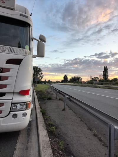 A white truck is parked on the shoulder of a highway. The truck is facing left in the frame, with its front end taking up the left side of the image. The truck is clean and appears to be in good condition. The highway is asphalt and has two lanes of traffic in each direction. The highway is bordered by a guardrail and a grassy shoulder. The sky is cloudy with some patches of pink and orange from the setting sun. The scene appears to be in a rural area, as there are trees and fields in the background. The location is identified as Saint-Aybert, France.