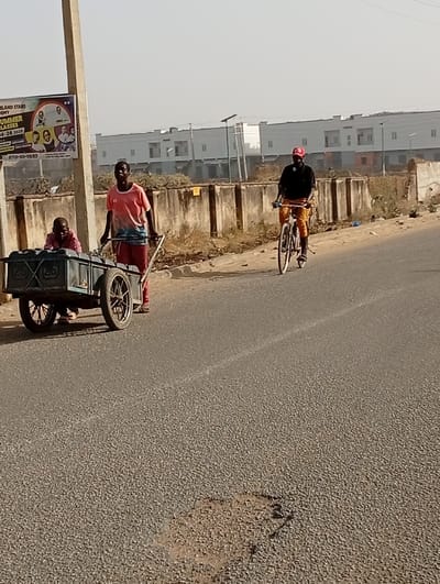 The image depicts a street scene in Gayawa, Nigeria, during daylight hours under a clear, bright sky. Three males are positioned on and alongside an asphalt road. On the left, a male wearing a pink and blue short-sleeved shirt with the number "10" on the chest pushes a two-wheeled, dark-colored metal handcart. Inside the handcart, a second male, wearing a red shirt, is seated low. To the right, a third male, wearing a black long-sleeved shirt, brown trousers, a red baseball cap, and a black object around his neck, rides a bicycle. All three individuals are moving in the same general direction, towards the left of the frame. The asphalt road surface exhibits minor wear and a prominent brown pothole in the foreground. A light-colored utility pole on the far left partially obscures a colorful billboard. The billboard displays text including "LAND STARS ACADEMY," "SUMMER CLASSES," "04-28-2023," and a phone number "0701-101-0593." A low, concrete barrier runs parallel to the road, behind which are piles of dry vegetation and several light-colored, multi-story buildings in the distance. Small, dark-colored street lights are visible along the distant barrier.