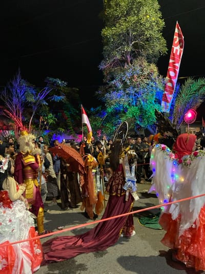 The image depicts a nighttime outdoor cultural parade or festival in Kedungbanteng, Indonesia. The scene is illuminated by artificial lights, including multicolored lights strung through trees and general ambient lighting.

In the immediate foreground, a person in a white and red multi-layered costume is partially visible on the left, beside another individual wearing elaborate maroon and gold warrior-like attire with a feathered or furred headpiece. Directly in front, a person in a long, dark maroon gown with a patterned bodice and an extensive train is visible from behind. This individual wears a large, light blue bow on their back, adorned with what appear to be paper currency notes. To their right, another participant is dressed in an orange-brown ensemble, carrying a decorative, open umbrella. Further right and slightly behind, a person in a red headscarf is seen from the back, wearing large, illuminated white wings adorned with small, colorful LED lights. A red and white rope barrier separates the costumed participants from the crowd.

In the mid-ground and background, numerous spectators and additional participants are visible. Many onlookers are gathered along the side of a paved street or pathway. Overhead, trees are illuminated with vibrant blue, green, and red lights. An Indonesian flag, red and white, is prominently displayed on a pole. To the right of the flag, a tall red banner with the partial word "INDONES" in white lettering is visible, possibly referring to "Indonesia." The ground is a dark, paved surface. The atmosphere suggests a community event with elaborate costumes and festive lighting.