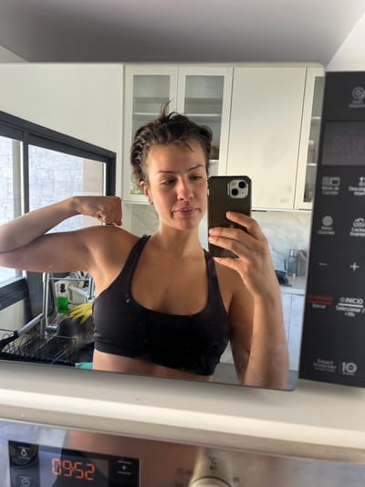 A female individual is captured in a mirror selfie within a modern kitchen setting in La Barra, Uruguay. The subject, with short, dark, wet hair, is wearing a black sports bra. She holds a dark-colored smartphone in her right hand to photograph her reflection while flexing her left arm, displaying her bicep. Her expression is neutral. The immediate foreground is the glossy white surface of a household appliance, likely an oven or washing machine, displaying a digital time of "09:52" and various control knobs/buttons. The background reveals a contemporary kitchen featuring white upper cabinets, some with glass inserts, above light-toned countertops. To the left, a large window with a black frame provides a view of an exterior wall constructed from light grey, textured stone blocks. Below this window, a sink area includes a black dish rack, a green liquid soap bottle, and yellow rubber gloves. To the right of the subject's reflection, the dark control panel of a microwave oven is visible, featuring white Spanish text such as "INICIO," "PARAR," "Borrar," "Menú Gourmet," and branding like "Smart Inverter," with a digital display showing "88:88." Natural light illuminates the scene from the left.