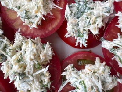 This is a close-up, top-down photograph of sliced tomatoes topped with a mixture of shredded ingredients and herbs. The tomatoes are bright red and appear to be sliced horizontally, revealing their inner texture and seeds. Each slice is generously topped with a creamy white mixture that includes finely shredded white ingredients, possibly cheese or a root vegetable, interspersed with green herbs, likely dill. Some darker, possibly shredded cheese bits are also visible within the topping. The overall impression is a visually appealing appetizer or side dish. The photograph is taken in a brightly lit indoor setting, with no discernible background elements to suggest a specific location within Подолино Ст, Russia.