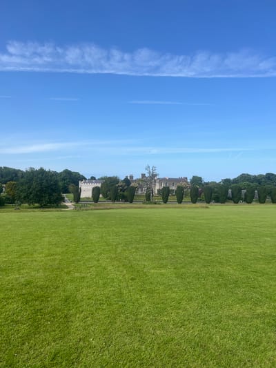 The image captures a wide, expansive view of a meticulously maintained green lawn in front of Ardgillan Castle in Ireland. The castle, a large, light-colored building with multiple windows and a distinct tower, is visible in the mid-ground, partially obscured by a line of tall, conical evergreen trees. To the left of the castle, a substantial portion of mature woodland can be seen. The foreground is dominated by the vast expanse of the lawn, which appears to be a park or open field. In the distance, a clear blue sky is dotted with wispy white clouds. Several picnic tables and benches are scattered across the lawn closer to the trees, suggesting a recreational area. The overall scene is one of serene, well-kept parkland and historical architecture under a bright sky.