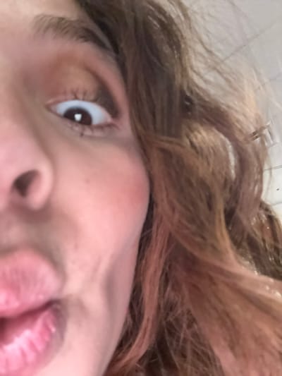 An extreme close-up focuses on a person's face from a low angle. The view includes the right eye, part of the nose, and mouth. The person has fair skin, brown hair, and dark eyelashes. The lips are pink and slightly parted. The eye looks upwards, showing the iris and a small glint of light reflecting off the pupil. The perspective and proximity create a distorted view of the facial features.