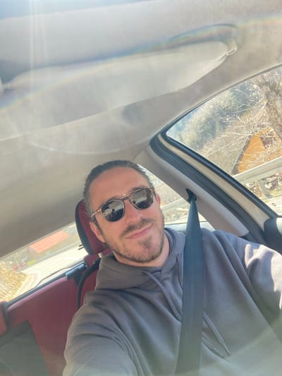 A man wearing sunglasses and a grey hooded sweatshirt sits in the driver's seat of a car. He is wearing a seatbelt. Outside the car window, trees and a building can be seen. The location is Lubnice, Montenegro.