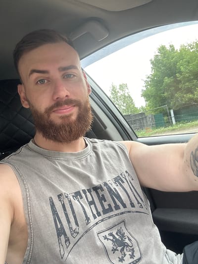 A man with short brown hair and a beard is sitting in the driver's seat of a car. He is wearing a gray sleeveless t-shirt with the word "AUTHENTICITY" printed on it in large letters. The car interior is visible, with a dark gray quilted seat. Through the side window, a fence and trees are visible, suggesting an outdoor, possibly rural, setting. The sky is overcast. The photo appears to be taken in the city of Yermakovskoye, Russia, based on the provided location context.