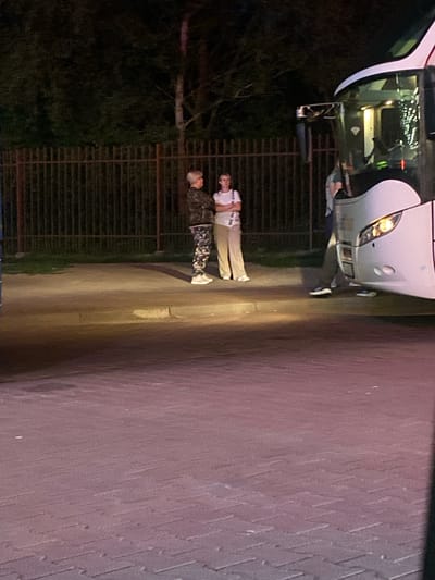 The image shows two individuals standing next to a white bus in what appears to be an outdoor setting at night. The bus is large and has its headlights on. The ground is paved with interlocking blocks, and a dark fence is visible behind the individuals. One individual is wearing a white t-shirt and beige pants, with their arms crossed. The other individual is wearing a camouflage-patterned jacket and pants. Both individuals are wearing white shoes. The lighting suggests it is nighttime, and the presence of the bus and paved area could indicate a bus station, parking lot, or a stop along a route. No specific emotions are discernible from their stances. There is no visible text in the image.