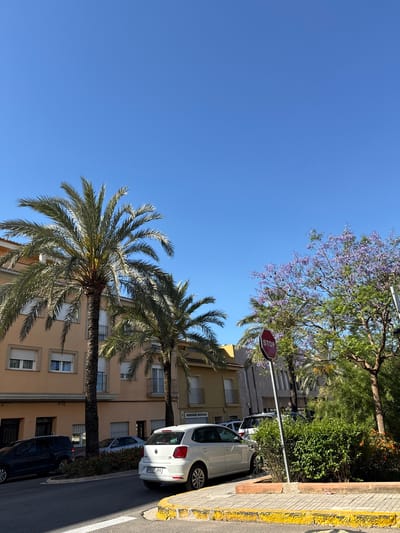 The image shows a street scene in l'Alcúdia de Crespins, Spain on a clear, sunny day. A blue sky occupies the upper left portion of the frame. Two tall palm trees stand adjacent to a multi-story beige building with windows and a balcony. A stop sign is visible further along the street, near a flowering tree with purple blossoms. Parked cars line the right side of the street, including a white Volkswagen. The pavement is asphalt with a yellow-painted curb.