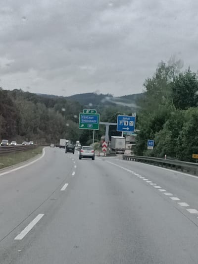 The image depicts a highway scene on a cloudy day. In the foreground, there is a two-lane road with white lane markings. Several cars are visible on the road, including a silver car in the right lane and other vehicles further ahead. On the right side of the road, there are trees and foliage. Overhead signs are present, indicating directions to "ČERČANY CHOCERADY" and a parking area with a "P" symbol. A blue sign with "40" is also visible, likely indicating a speed limit. In the background, rolling hills covered in trees are seen under a grey sky. The scene appears to be a section of highway in or near Vranov, Czechia, given the road signs.