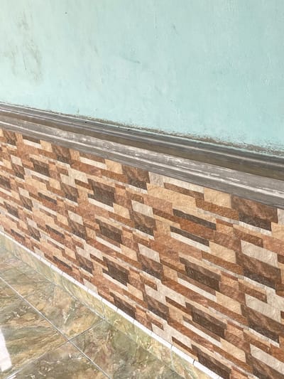 The image shows a close-up view of a textured surface, possibly a wall or ceiling, with a tiled border. The majority of the image is dominated by a light blue, textured surface that appears to be painted or stuccoed. To the right of this surface, there is a decorative border with multiple ribbed layers, transitioning into a patterned tile section. These tiles are rectangular and arranged in a staggered pattern, featuring a mosaic of brown, beige, and white tones. Further to the right, the pattern changes to larger, marbled tiles in shades of green and brown, also arranged in a grid.
The lighting suggests it is daytime, possibly indoors or in a shaded outdoor area given the even illumination and lack of harsh shadows. There are no people or clear indicators of specific activities taking place. The setting appears to be a finished interior or exterior of a building, characterized by its architectural finishes. The location context provided is Sekar Mawar, Indonesia, but there are no specific visual cues within the image that confirm this location. No visible text is present in the image.