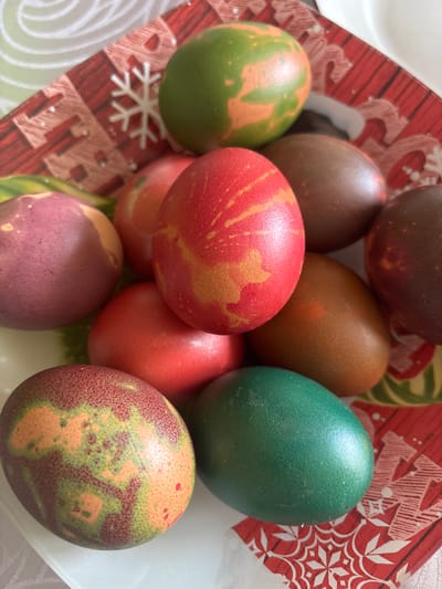 The image shows a collection of dyed Easter eggs in a bowl with a festive, holiday-themed design. The eggs are various colors, including red with golden streaks, green, brown, and a purplish-red with mottled patterns. The bowl appears to be located indoors, based on the lighting and the presence of a tabletop beneath the bowl. The exact location is Klimentovo, Bulgaria.