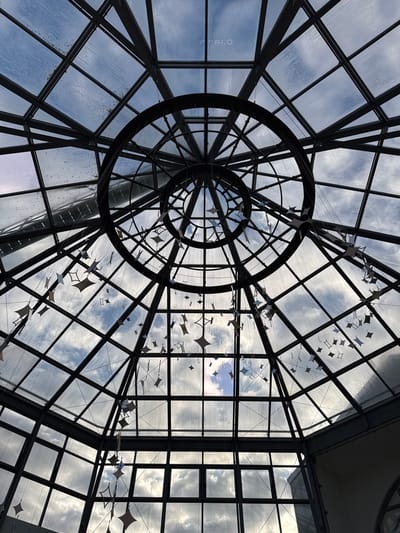 The image depicts an upward view of a large, domed glass skylight or ceiling structure located within a building in Serris, France. The dome is constructed with a dark, metallic grid framework forming numerous polygonal glass panes. At the apex, a concentric circular metal structure with two prominent rings is visible, from which numerous star-shaped or diamond-shaped decorative elements are suspended by thin lines, appearing in various shades of light grey and white. Through the glass, a partially cloudy sky is visible, featuring patches of blue sky interspersed with white and grey clouds. Some glass panes exhibit signs of condensation or dirt on their exterior surfaces. No individuals are present in the frame. The scene is illuminated by natural light filtering through the glass.