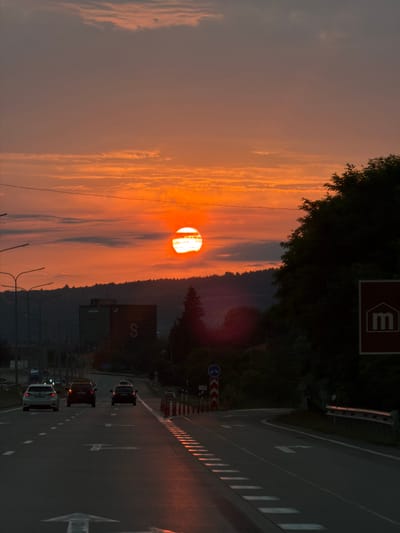The image depicts a road scene at sunset, with the sun prominently visible in the orange sky. The road appears to be a highway or a major thoroughfare, with multiple lanes and directional arrows painted on the asphalt. Several cars are driving on the road, with their headlights on, suggesting it is dusk or early evening. On the right side of the road, there is a dark green tree line and a red sign with a white "m" inside a house-like shape. In the background, there are some buildings, including what appears to be a large, dark structure with the letter "S" visible on it, and a landscape of hills covered in trees. The sky is filled with streaks of orange and pink clouds, indicative of a sunset. The overall scene is bathed in the warm, diffused light of the setting sun. There is no discernible indication of the location being in Kveda Simoneti, Georgia.