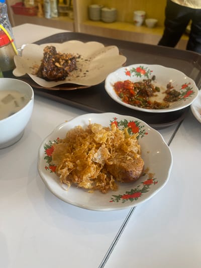 An overhead shot shows a white table laden with Indonesian food. On the left, a white bowl contains a light-colored soup with green garnish. In the center, a plate holds fried, crispy food that appears to be egg or a similar batter. To the right, another small white bowl contains a spicy red chili and vegetable condiment. Further back, a larger plate is adorned with a dark glazed meat dish, possibly chicken or beef, topped with fried shallots, served on a piece of textured brown paper. The food is arranged on a dark brown tray, suggesting it was served from a restaurant. The background shows shelves with stacked plates and bottles, indicating a dining establishment in Medan, Indonesia.