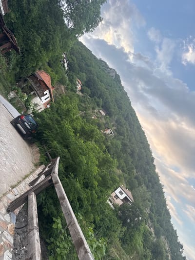 The image depicts a scenic hillside village, likely in Delchevo, Bulgaria, bathed in the soft light of late afternoon or early evening. The sky above is a dramatic mix of blue and grey clouds, with patches of warm sunlight breaking through, suggesting dynamic weather. The scene is dominated by a steep, densely forested slope, dotted with a scattering of traditional houses. These homes, built with stone and wood, feature characteristic red-tiled roofs and balconies, blending harmoniously with the natural landscape. Some houses appear well-maintained, while others show signs of age and disrepair, hinting at a community with a rich history.
In the foreground, a cobblestone path winds its way up the hill, bordered by a rustic wooden fence and a stone wall. A dark-colored SUV is parked on the path, its license plate bearing the text "E 293 MH". The overall impression is one of tranquility and rustic charm, with the natural beauty of the mountainside juxtaposed against the human element of the village. The dense greenery suggests a fertile environment, possibly late spring or summer. No people are visible, contributing to a sense of peaceful solitude.