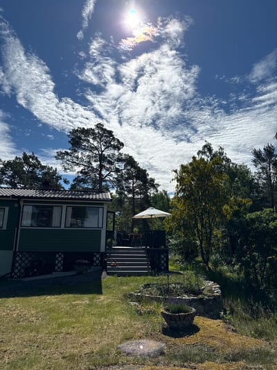 This is a photograph taken in Stavsnäs, Sweden, on a sunny day with a bright blue sky filled with white, wispy clouds. The sun is high in the sky, with lens flare visible. The scene depicts a green house with a dark-tiled roof, situated in a grassy yard surrounded by trees. A wooden deck with an umbrella and outdoor furniture is attached to the side of the house, accessible by a set of stairs. In the foreground, there is a garden bed with various plants and a decorative stone pot containing lavender. The overall environment suggests a peaceful, rural setting.