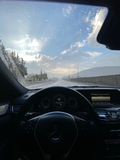 An interior view from a Mercedes-Benz car presents a rain-streaked windshield. Outside, a road stretches toward distant mountains under a sky filled with sunlit clouds. The dashboard gauges display "40" and "11.5°C". The steering wheel prominently features the Mercedes logo. The central console and infotainment screen are partially visible. The perspective is slightly tilted.