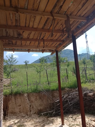 The image depicts an unfinished structure with a wooden roof supported by reddish-brown metal beams. The roof is constructed from rough-hewn wooden planks. Beyond the structure, a grassy hillside is visible, dotted with small trees and purple flowers. In the background, a large mountain range rises against a hazy blue sky with scattered clouds. A prominent power line tower stands to the right of the frame. The ground in the foreground consists of sandy soil with a pile of branches to the right. A section of a brick wall is visible on the left side of the image. The scene suggests an outdoor, possibly rural, location in or near Kerben, Kyrgyzstan, characterized by construction, natural landscape, and infrastructure elements.