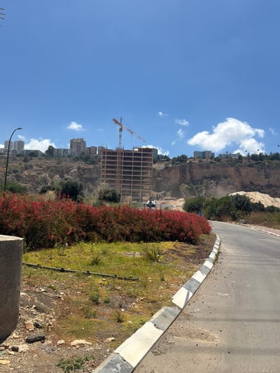 The image captures a construction site with a large building under development, dwarfed by a cliff face. A prominent construction crane, painted in red and white, stands tall against a bright blue sky dotted with white clouds. Several other buildings, presumably residential, are visible nestled into the slopes of the cliff, suggesting a city or town built into a hilly or mountainous terrain.
In the foreground, a paved road curves along the edge of the scene, bordered by a strip of green grass and vibrant red flowering bushes. The grass is patchy in places, with visible soil and scattered rocks, indicating a somewhat arid or unkempt landscape. A concrete curb with alternating white and grey sections separates the road from the grassy verge.
The overall scene suggests a sunny day, likely late morning or early afternoon, given the brightness and the clear blue sky. The presence of cable cars on the cliff face hints at a location with significant elevation changes, possibly a city known for its dramatic topography. The construction activity implies ongoing development and progress. No people are visible in the frame, and no text is present. The location context of Nesher, Israel, aligns with the visual cues of a Mediterranean climate and possibly a city built on challenging terrain.